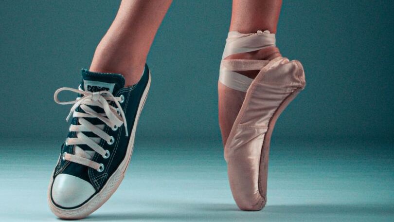 Ein Fu&szlig; in einem Ballettschuh und ein Fu&szlig; in einem Turnschuh heben sich in der Luft vor einem neutralen Hintergrund.