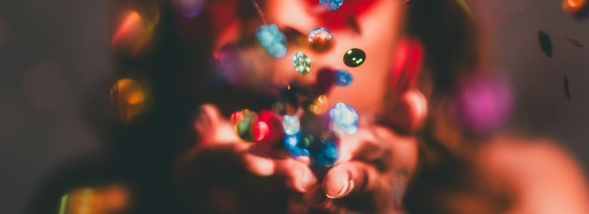 Frau bläst bunten Konfetti aus ihren Händen, während sie in die Kamera schaut, mit verschwommener Hintergrundbeleuchtung.