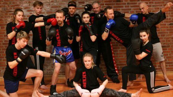 Gruppe von zw&ouml;lf Kampfsportlern in Schwarz, posierend mit Boxhandschuhen vor einer Ziegelsteinwand in einem Trainingsraum.