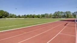 Luftaufnahme eines leeren Sportplatzes mit einer roten Laufbahn und gr&uuml;nem Rasen im Hintergrund unter blauem Himmel.