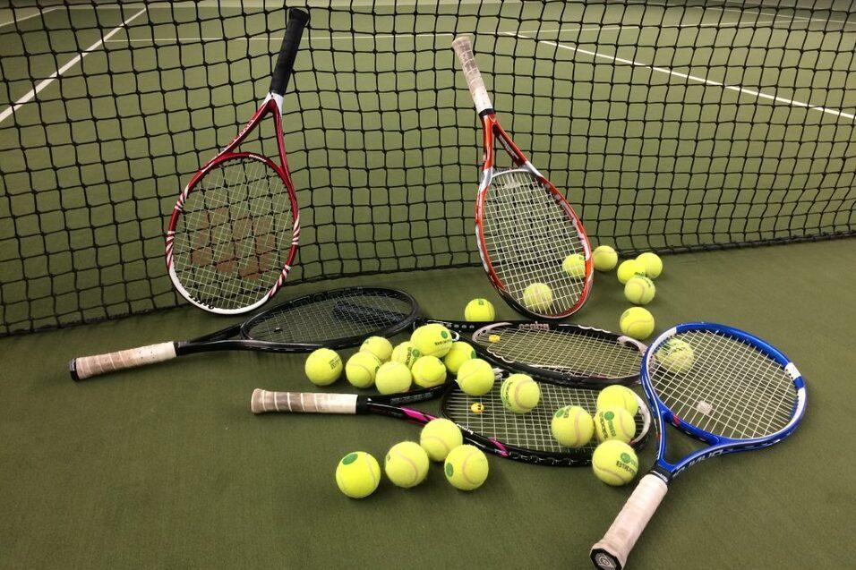 Drei Tennisschl&auml;ger und zahlreiche gelbe Tennisb&auml;lle liegen auf einem Tennisplatz hinter einem Netz.