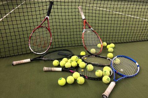 Drei Tennisschläger und zahlreiche gelbe Tennisbälle liegen auf einem Tennisplatz hinter einem Netz.