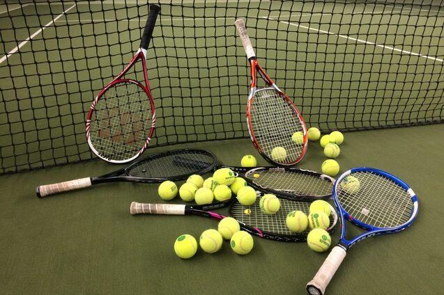 Drei Tennisschl&auml;ger und zahlreiche gelbe Tennisb&auml;lle liegen auf einem Tennisplatz hinter einem Netz.
