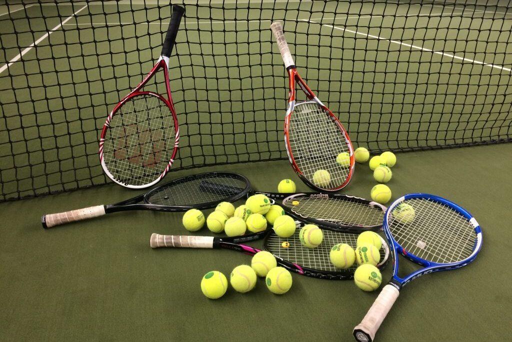 Drei Tennisschl&auml;ger und zahlreiche gelbe Tennisb&auml;lle liegen auf einem Tennisplatz hinter einem Netz.