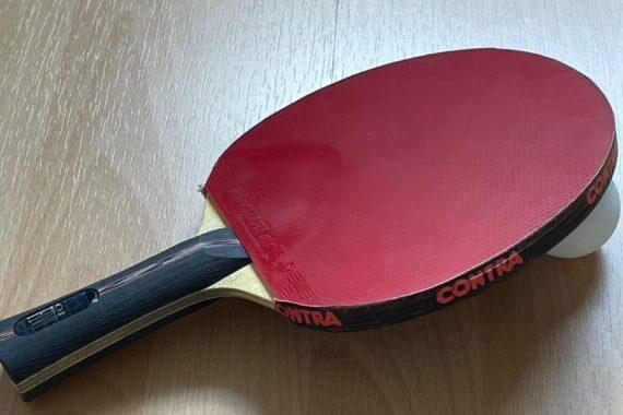 Nahaufnahme eines roten Tischtennisschl&auml;gers mit Ball auf einer Holzoberfl&auml;che.