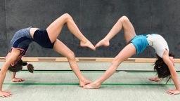 Zwei M&auml;dchen f&uuml;hren eine Br&uuml;cke in entgegengesetzter Richtung durch, verbunden durch ihre Beine, auf einem Sportboden.