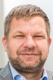 Portr&auml;t eines Mannes mit kurzem Haar, Bart und grauem Blazer, der direkt in die Kamera blickt.