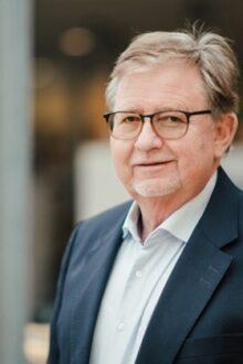 Portr&auml;t eines &auml;lteren Mannes mit Brille, l&auml;chelnd in einem modernen, hellen Raum. Er tr&auml;gt einen dunklen Anzug und ein helles Hemd.