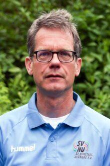 Portr&auml;t eines Mannes mit Brille, der ein hellblaues Polo-Shirt tr&auml;gt, vor einem gr&uuml;nen Hintergrund.