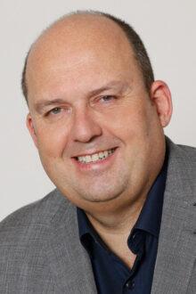 Portr&auml;t eines l&auml;chelnden Mannes mit glattem Kopf, in grauem Anzug und dunkelblauer Hemd, vor hellem Hintergrund.