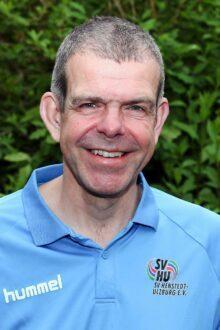 Portr&auml;t eines l&auml;chelnden Mannes in hellblauem Shirt vor gr&uuml;ner, &uuml;ppiger Hintergrundvegetation.