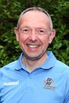 Portr&auml;t eines l&auml;chelnden Mannes in blauer Sportbekleidung vor gr&uuml;nem Hintergrund mit Pflanzen.