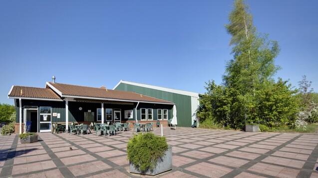 Gr&uuml;nes Geb&auml;ude mit Terrasse, abgegrenzt von Pflanzen und Tischen im Freien, unter blauem Himmel.