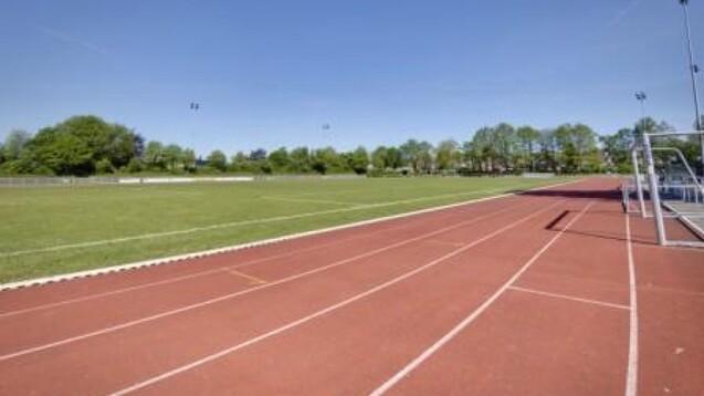 Leere Sportanlage mit rotem Laufbahn und gr&uuml;nem Fu&szlig;ballfeld unter blauem Himmel und B&auml;umen im Hintergrund.