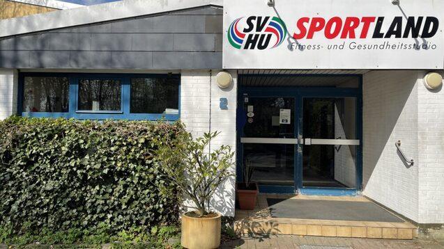 Eingang zum Fitnessstudio "SV Hu Sportland" mit blauen Fenstern und gr&uuml;ner Bepflanzung vor der wei&szlig;en Wand.