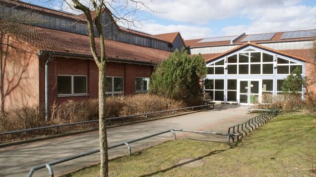 Eingang eines Geb&auml;udes mit Glasfront, umgeben von B&uuml;schen und einem gepflasterten Weg. Teilweise bew&ouml;lkter Himmel.