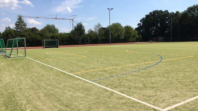 Gr&uuml;nes Kunstrasenfeld mit zwei Fu&szlig;balltoren und Markierungen, umgeben von B&auml;umen und einem Kran im Hintergrund.