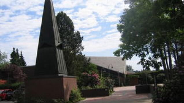 Sicht auf einen hohen, spitzen Obelisk vor einem Geb&auml;ude, umgeben von B&auml;umen und bl&uuml;henden Str&auml;uchern.