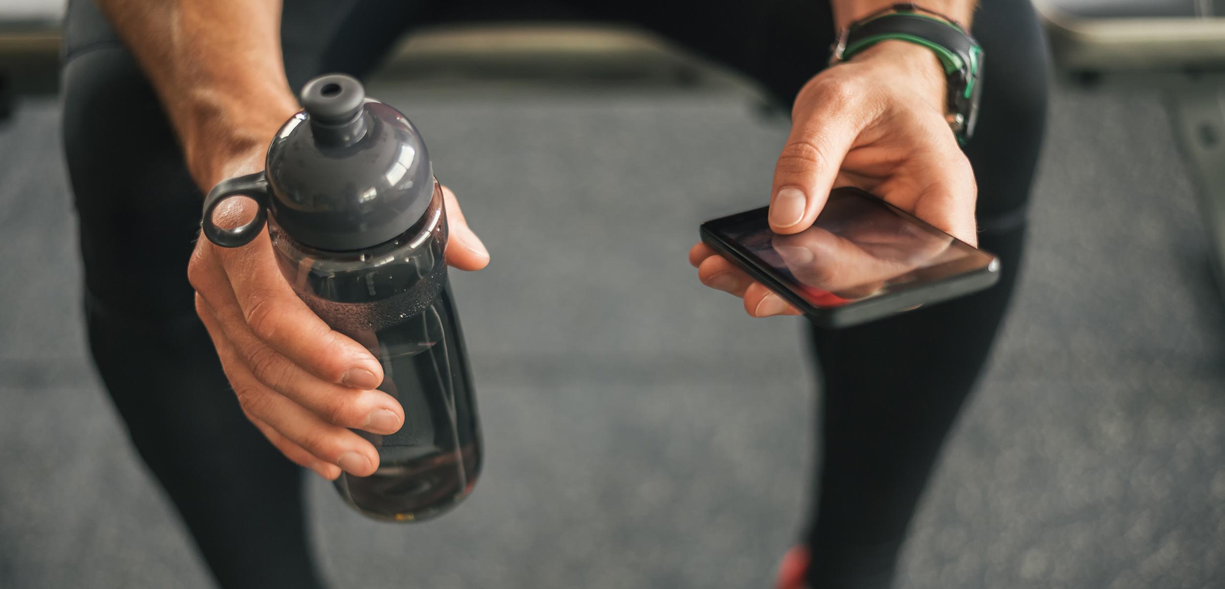 Eine Hand h&auml;lt eine Trinkflasche, w&auml;hrend die andere ein Smartphone in einem Fitnessstudio ber&uuml;hrt.