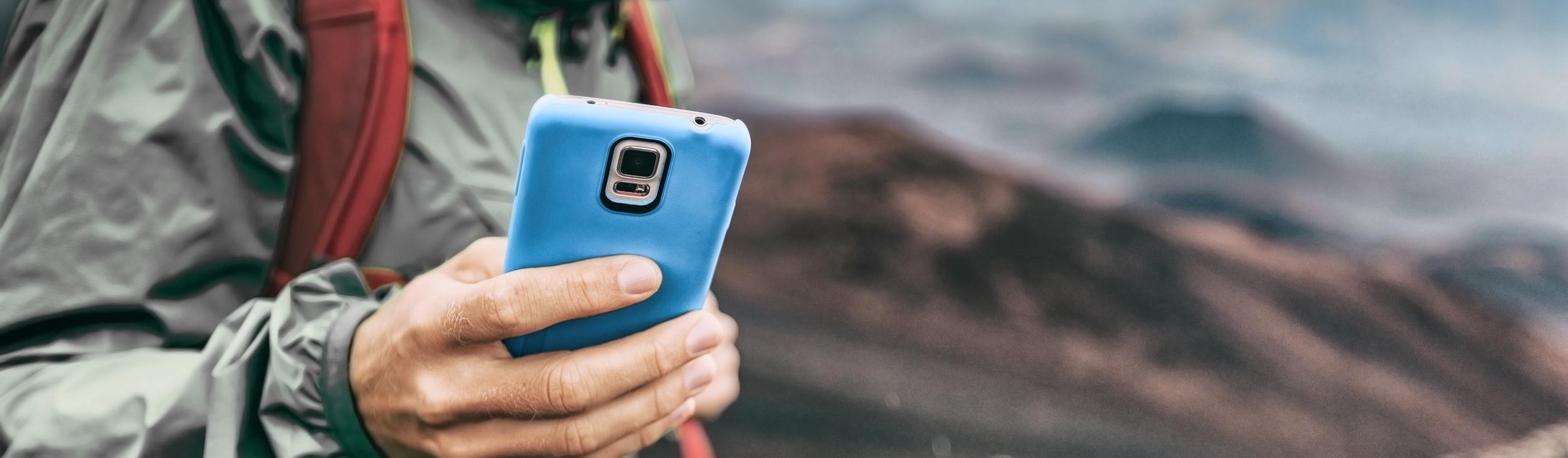 Ein Wanderer in Outdoor-Bekleidung h&auml;lt ein Smartphone in der Hand, w&auml;hrend er die Berglandschaft betrachtet.