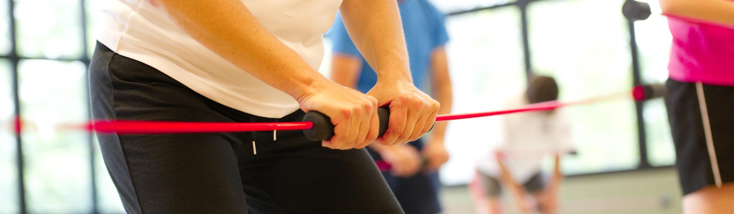 Nahaufnahme einer Person mit einem Widerstandsband im Fitnessstudio, umgeben von weiteren trainierenden Personen.