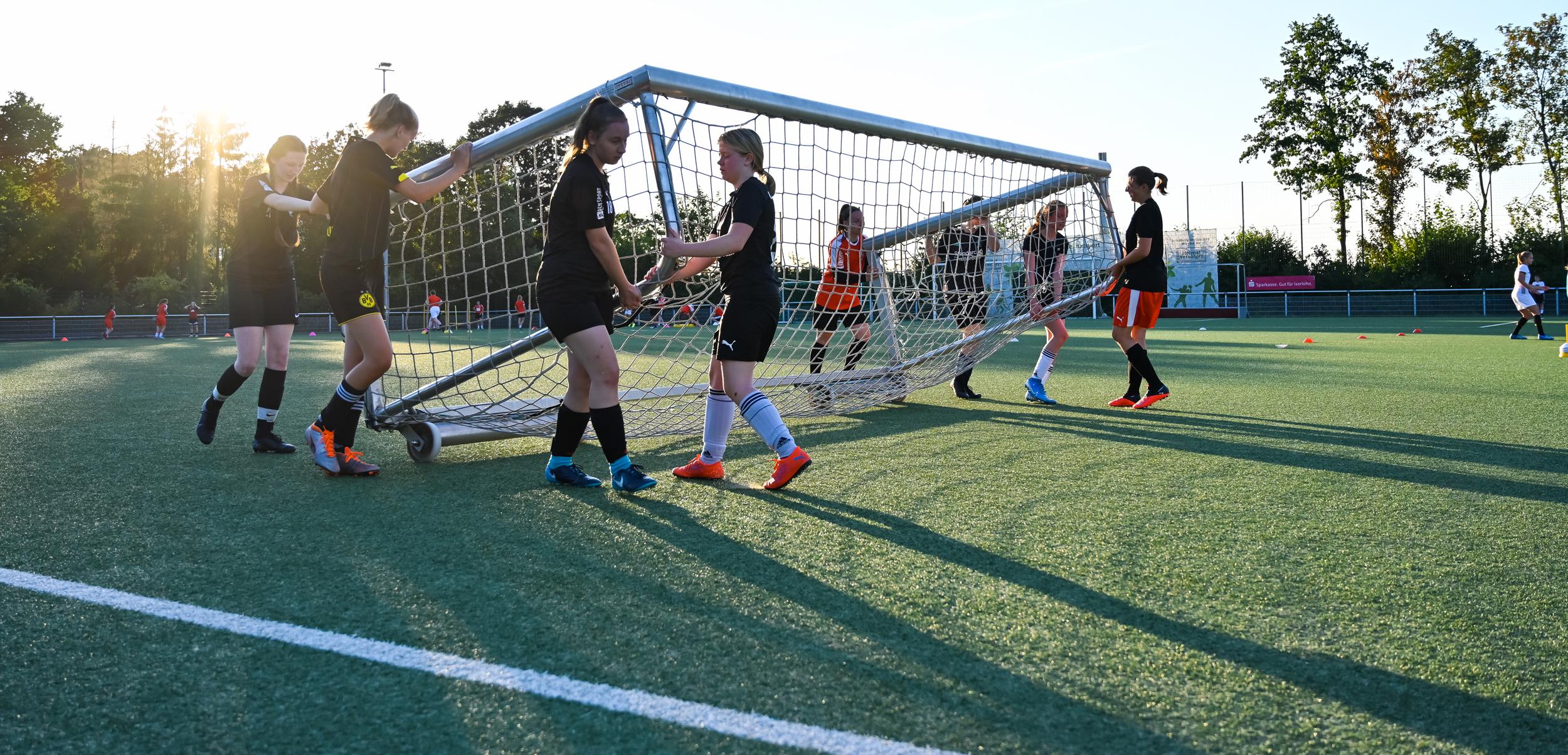 Mannschaft von Fußballspielerinnen hebt ein Tor auf einem Kunstrasenplatz bei Sonnenuntergang an.