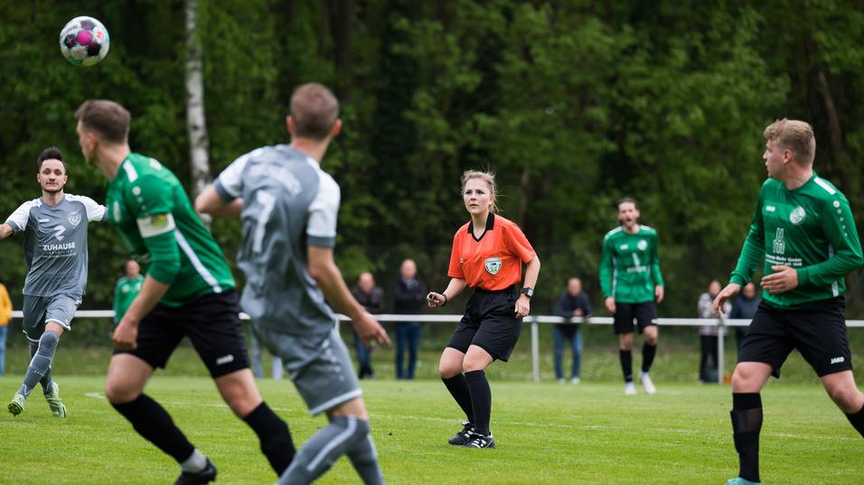 Fußballspiel mit einem Ball in der Luft und einer Schiedsrichterin in orange, umgeben von spielenden Spielern und Zuschauern.