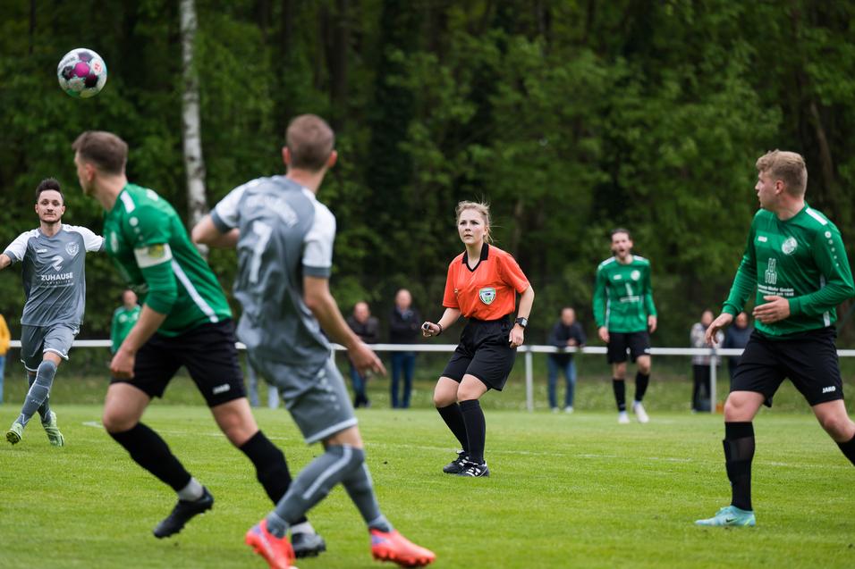 Fußballspiel mit einem Ball in der Luft und einer Schiedsrichterin in orange, umgeben von spielenden Spielern und Zuschauern.