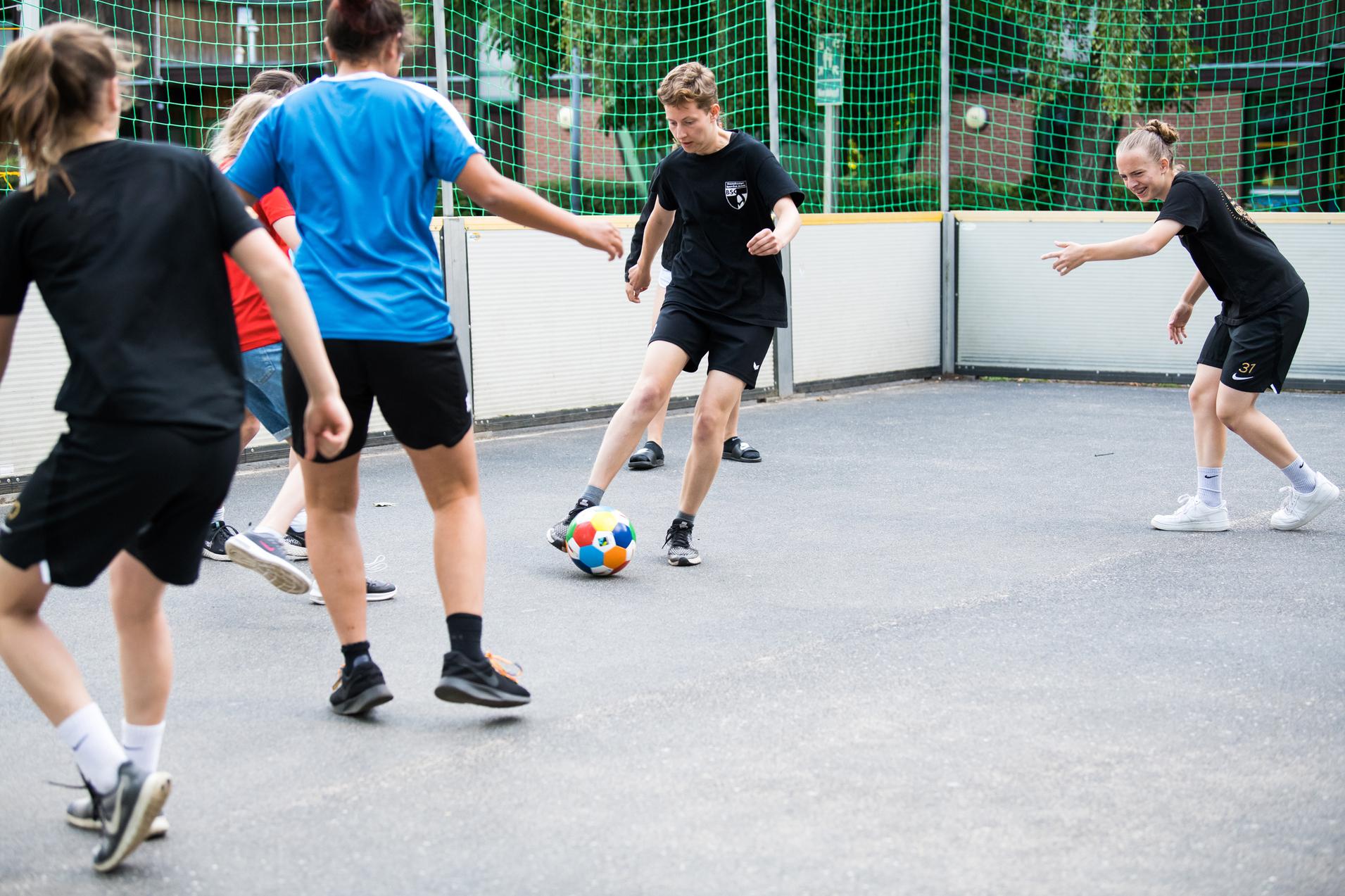 Gruppe von Jugendlichen spielt Fußball in einem umschlossenen Sportbereich mit Netz und Gummiuntergrund.