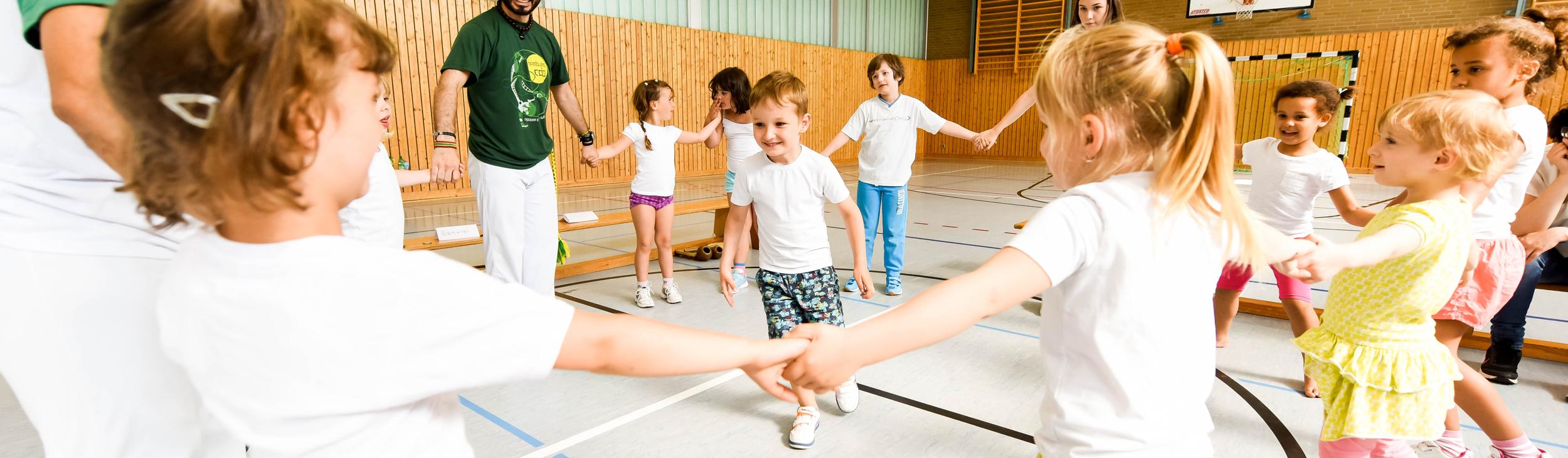 Gruppe von Kindern in T-Shirts und Erwachsenen in einer Turnhalle, die gemeinsam einen Kreis bilden und spielen.