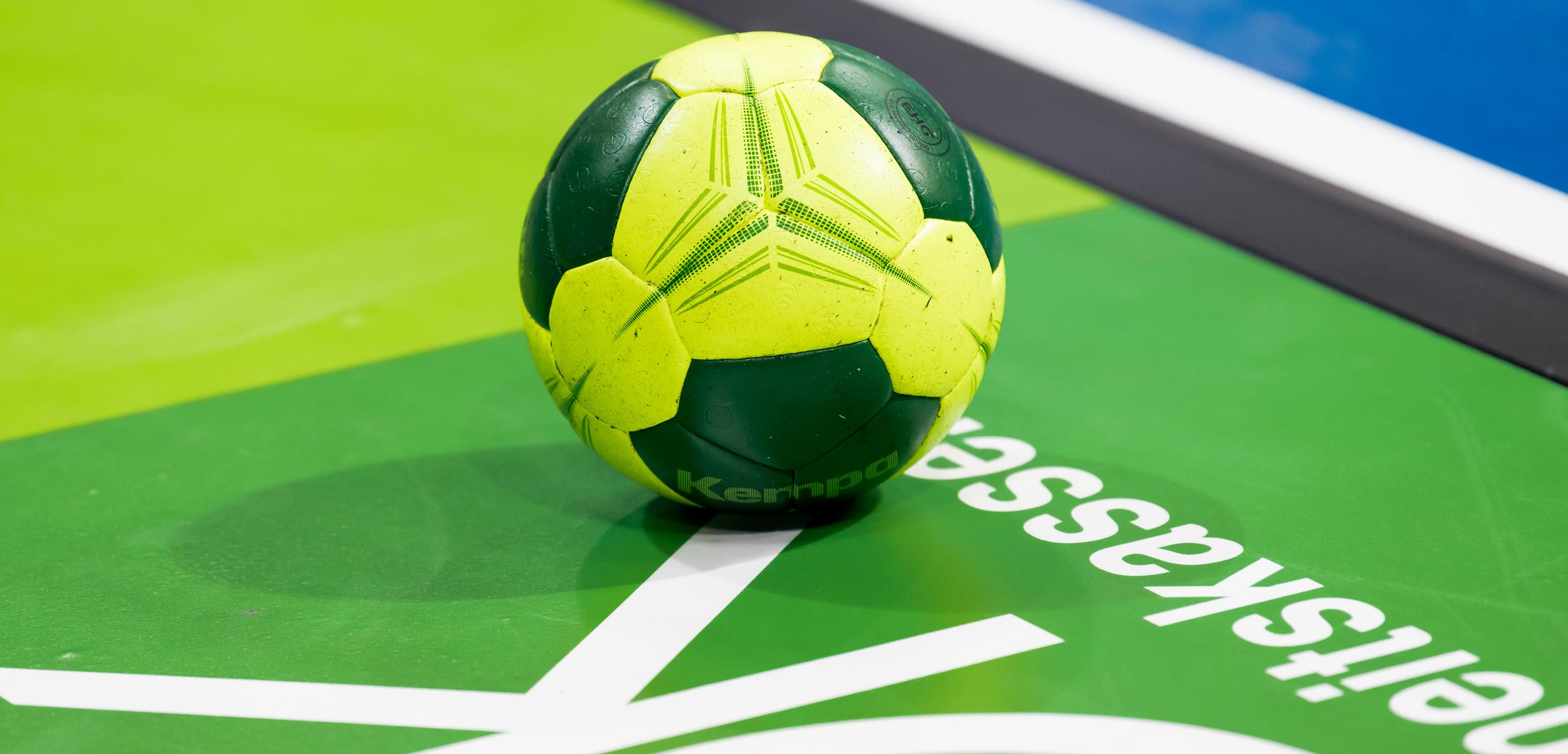 Grüner und gelber Handball liegt auf einer grünen Spielfläche mit weißem Schriftzug.