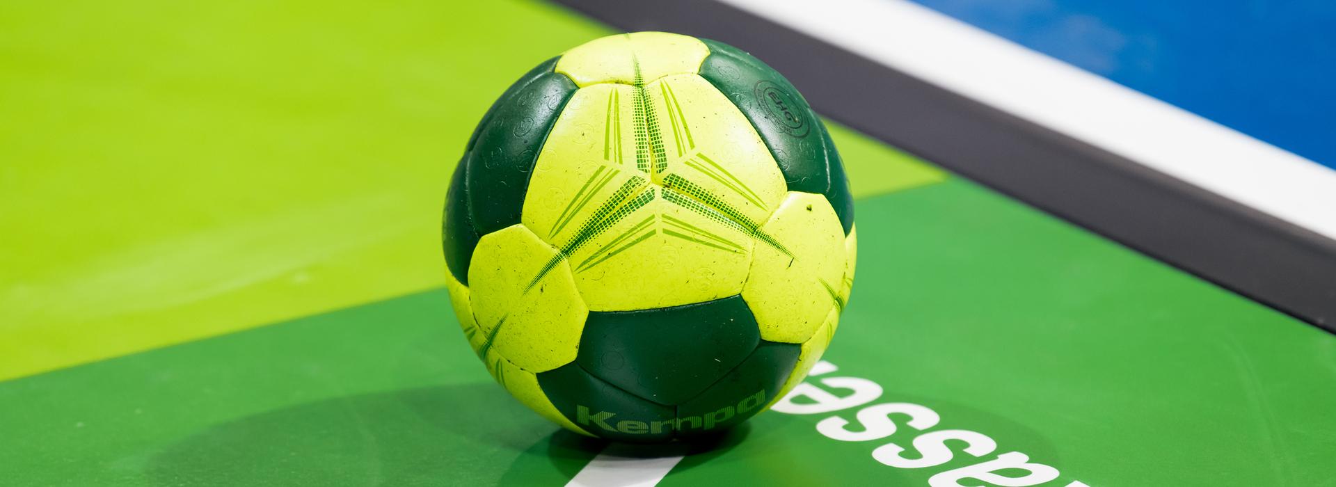 Grüner und gelber Handball liegt auf einer grünen Spielfläche mit weißem Schriftzug.