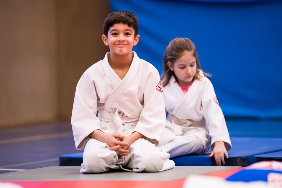 Zwei Kinder in weißen Judoanzügen sitzen auf einer Matte in einem Judoraum, der Hintergrund ist blau.