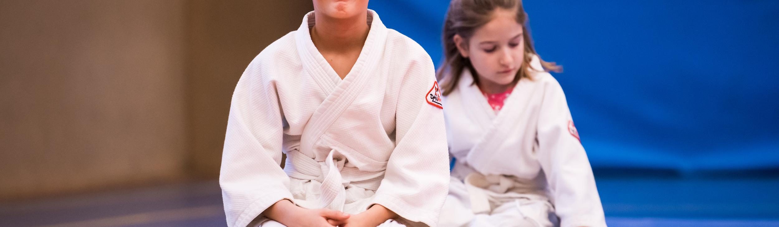 Zwei Kinder in weißen Judoanzügen sitzen auf einer Matte in einem Judoraum, der Hintergrund ist blau.