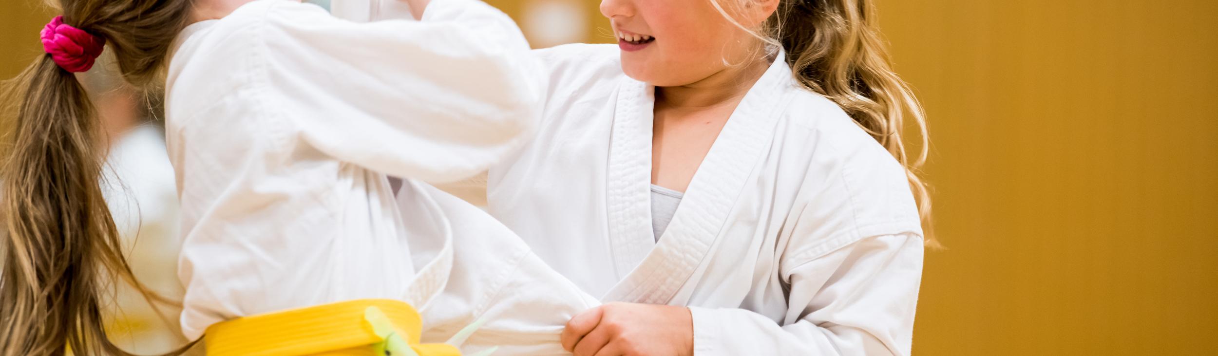 Zwei Mädchen in weißen Karateanzügen mit gelben Gurten üben freundschaftlich im Trainingsraum miteinander.