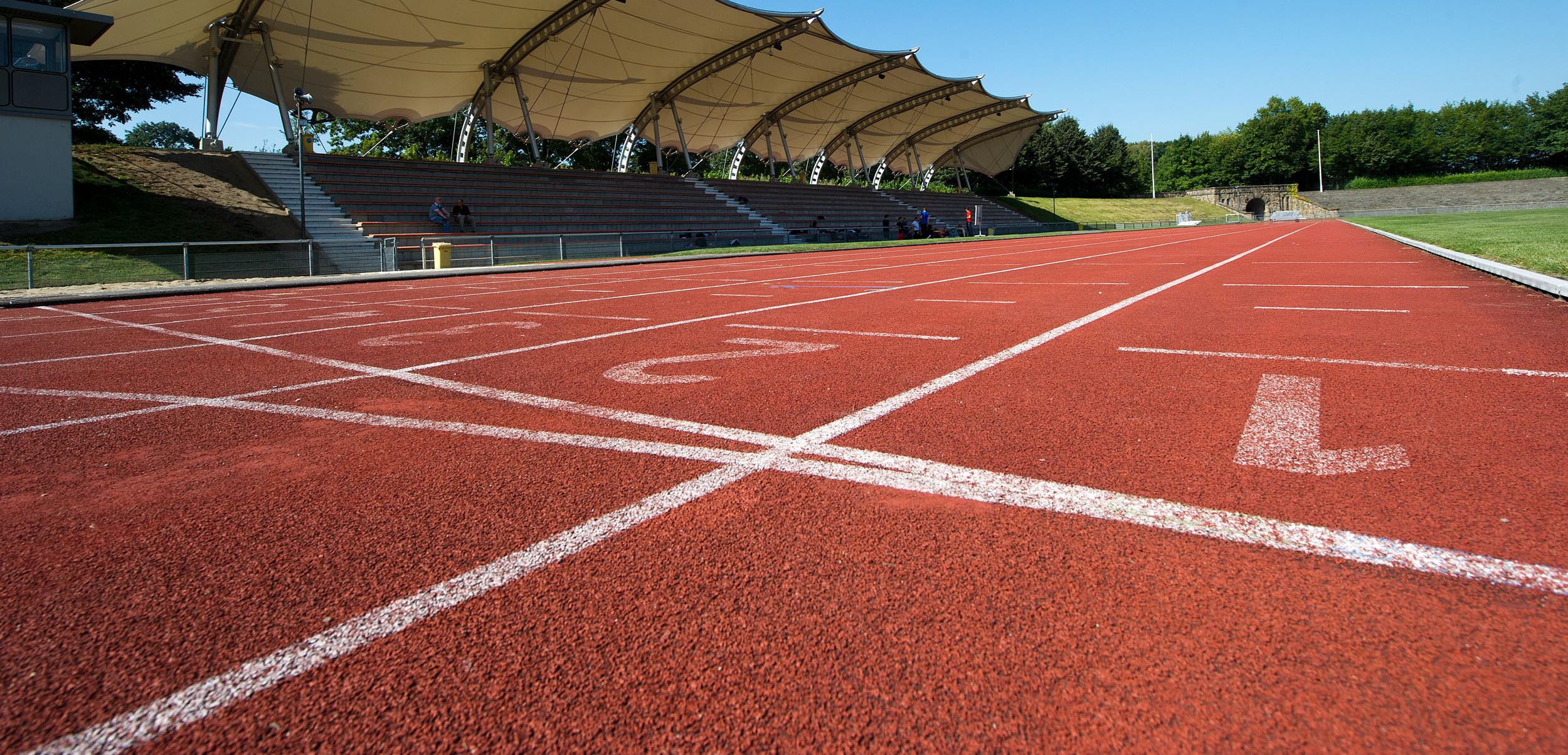 Nahaufnahme einer roten Laufbahn mit Startlinien und einem überdachten Zuschauerbereich im Hintergrund.