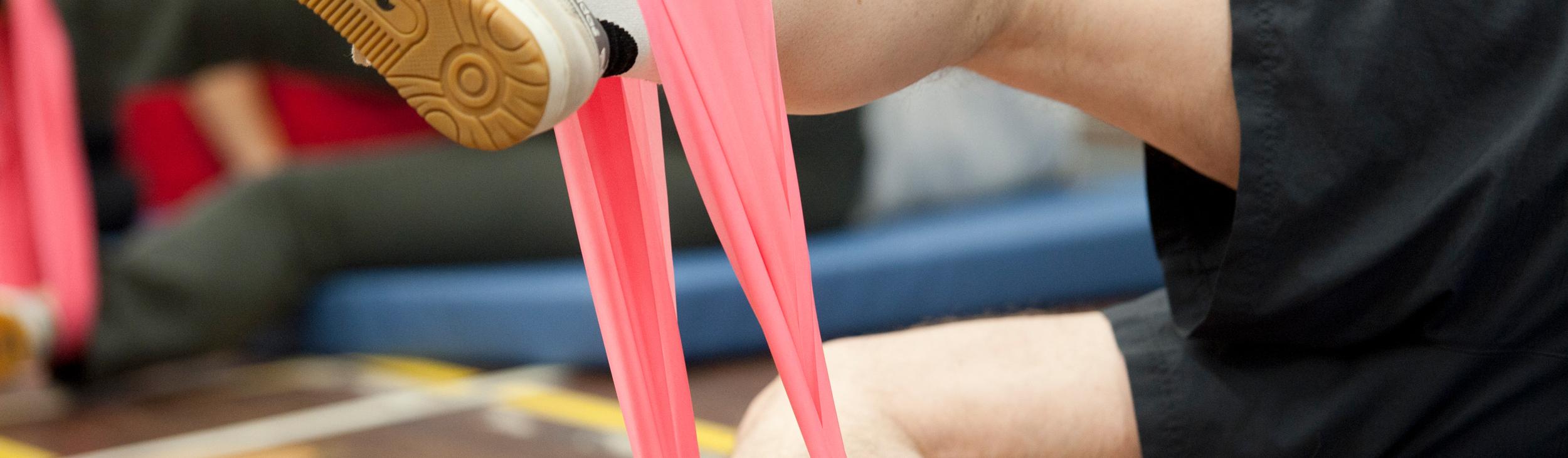 Bein eines Sportlers mit pinkem Widerstandsband, auf einer Sportmatte, im Hintergrund weitere Teilnehmer.