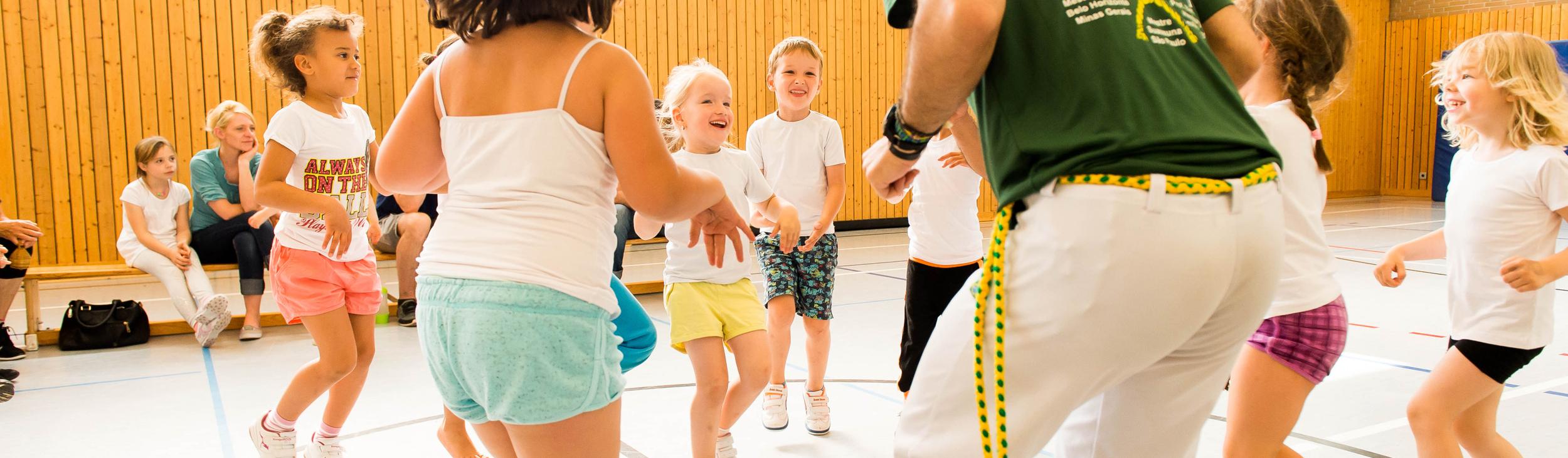 Gruppe von Kindern tanzt fröhlich in einer Turnhalle, geleitet von einem Trainer in grünem T-Shirt.