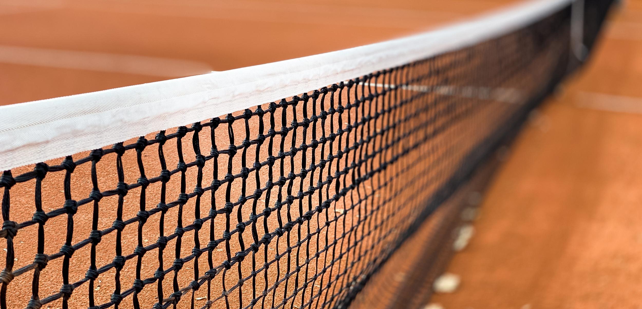 Nahaufnahme eines Tennisnetzes auf einem roten Sandplatz, Hintergrund unscharf mit grünen Bäumen.