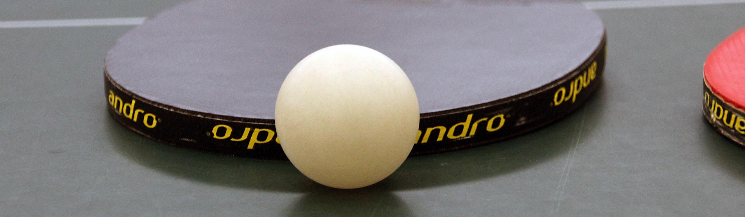 Nahaufnahme eines Tischtennisschlägers mit weißem Ball auf einer grünen Tischtennisplatte.