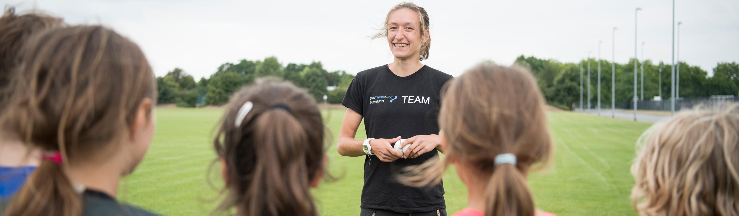 Trainerin spricht mit jungen Sportlerinnen auf einem grünen Fußballfeld unter bewölkten Himmel.