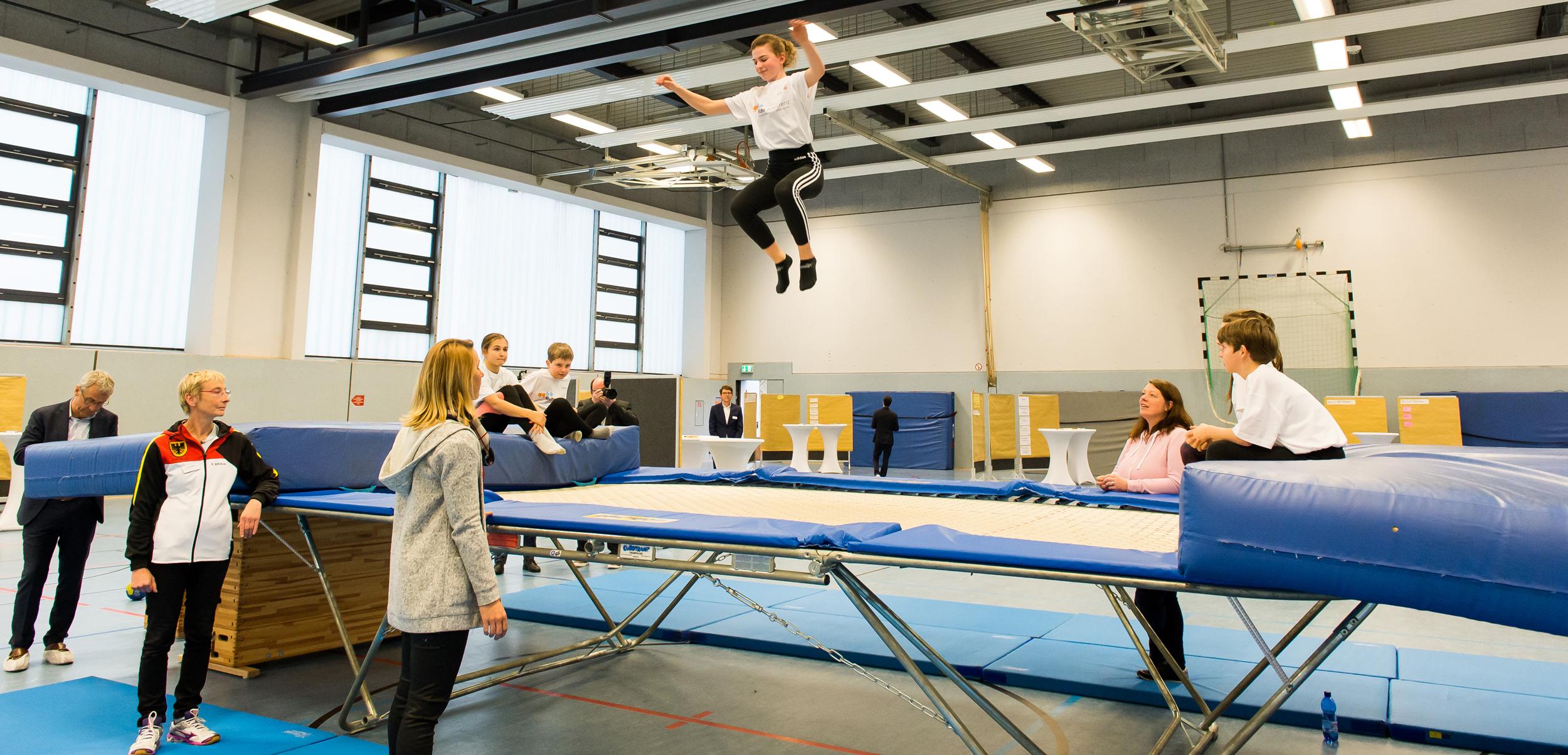 Junge springt auf einem Trampolin, während Zuschauer und Trainer im Hintergrund beobachten. Sporthalle mit hellem Licht.